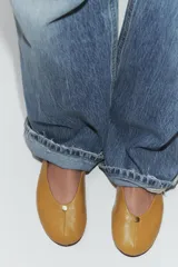 Zapato tipo bailarina plana de piel en color tostado, con pespuntes en el cuerpo, detalle metálico en la parte delantera y acabado en punta redonda.