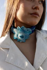 Gargantilla tejida a mano con hilo azul, adornada con una flor central de tela y detalles de pedrería.