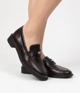 Mocasines de poliuretano color marrón oscuro, tipo penny loafer, con costuras visibles alrededor de la puntera y detalle de adorno metálico dorado en el empeine. Poseen suela baja con pequeño tacón cuadrado.