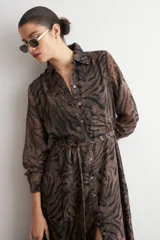 Vestido camisero largo con estampado animal print tipo cebra, en tonos marrón y negro, con cuello camisero, mangas largas y cierre frontal con botones. Incluye un cinturón fino de cordón negro.