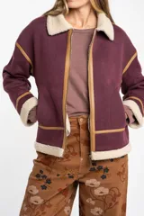 Campera tipo gamulán de color bordó, con interior y cuello de corderito sintético en tono crema. Presenta detalles de ribetes en cuero sintético color suela, cierre frontal metálico y bolsillos laterales.