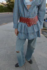 Vestido corto de manga larga con cuello solapa y cierre frontal de botones a presión. Presenta un diseño a cuadros en tonos azul y marrón, cintura ajustable con cinturón de hebilla y falda con volantes en el bajo.