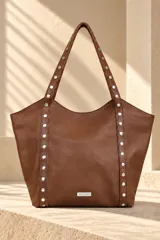 Cartera tipo tote de gamuza color azul marino, con asas dobles de cuero marrón texturado y detalles de tachas metálicas plateadas. Cuenta con una placa metálica frontal con el logo de la marca Pecarí.