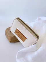 Billetera pequeña de cuero texturizado color crudo, con cierre metálico perimetral y parche frontal de cuero color suela con el logo de la marca grabado.