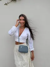 Bandolera pequeña de diseño rectangular en color beige, confeccionada en cuero sintético. Presenta una correa larga ajustable del mismo material y cierre superior con cremallera. Incluye el logo de la marca grabado en la parte frontal.