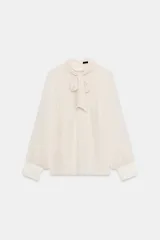 Camisa cruda de manga larga con textura plumeti, cuello redondo y lazada en el mismo tejido. Cuenta con mangas abullonadas terminadas en puños anchos y cierre en la espalda con abertura y botones.