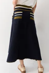 Falda midi de lana en color marrón, con diseño acampanado y tejido acanalado. Presenta una cintura elástica con cordón de ajuste, detalles de rayas en tonos beige y blanco en la parte superior, y costuras decorativas en el ruedo.