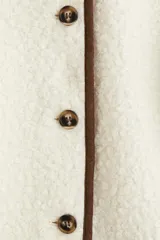 Saco corto de tejido sherpa color crema, con cuello redondo y cierre frontal de botones marrones. Presenta ribetes de tela marrón en el escote y la tapeta frontal.