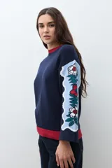 Sweater de punto color azul marino con diseño jacquard floral en las mangas y detalle de cuello redondo en color rojo. Presenta un corte holgado y mangas tres cuartos.