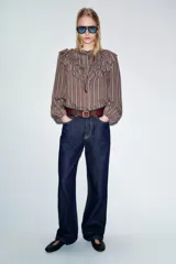 Camisa de manga larga con cuello subido, cierre frontal con botones y detalle de volantes en el pecho. Presenta un diseño a rayas verticales y puños elásticos.