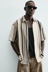 Camisa de manga corta con calce relaxed fit, confeccionada en tejido estructurado con algodón. Presenta un diseño de rayas verticales en tonos beige, negro y blanco, cuello solapa, cierre frontal con botones y aberturas laterales en el bajo.