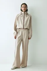 Campera rompevientos de color beige, modelo oversized con cuello alto y capucha con cordón ajustable. Cuenta con cierre frontal marrón, mangas raglán con puños elásticos, bolsillos laterales y detalles de ribetes en tono marrón oscuro.