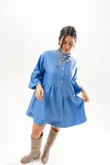 Vestido corto azul, estilo camisero, con cuello camisero, cierre frontal con botones y mangas acampanadas con volados. Presenta un diseño holgado con cortes fruncidos en el cuerpo.