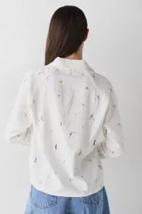 Camisa de manga larga color blanco con cuello polo y cierre frontal parcial. Presenta un estampado integral con motivos deportivos y un detalle bordado en el pecho.