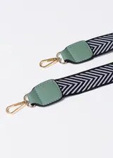 Bandolera verde agua con textura geométrica, cierre superior y correa de tela estampada en blanco y negro. Incluye charm colgante con forma de gato.