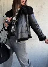 Campera tipo gamulán de cuero sintético negro con interior y detalles de cuello y puños en piel sintética negra. Presenta cierre frontal, correas ajustables en los puños y diseño de corte clásico.