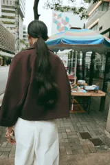 Trench corto de color marrón oscuro, con cuello de solapa, cierre frontal cruzado con botones y mangas tres cuartos con detalle de vuelta, trabilla y botón. Cuenta con bolsillos delanteros de vivo.