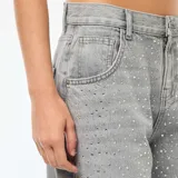 Jean de corte recto y tiro alto, color gris claro, con detalle de strass aplicado en toda la parte frontal. Cuenta con cierre de botón y cremallera, y bolsillos laterales y traseros.