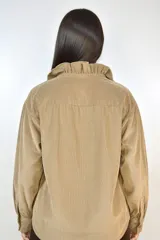 Camisa de pana fina color beige, con volados decorativos en los hombros y a lo largo de la botonadura frontal. Presenta cuello clásico, mangas largas con puños y cierre mediante botones dorados.