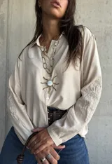Camisa de corte holgado en color beige, con cuello clásico, cierre frontal de botones y mangas largas que presentan un delicado bordado tonal en los antebrazos.