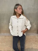 Saco tejido de punto con diseño de ochos y trenzas, color crudo. Presenta cuello camisero, cierre frontal con botones joya y mangas holgadas con puños acanalados.