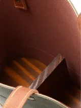 Matera de diseño vertical con cuerpo en cuero verde oscuro y base en cuero color suela. Presenta correas largas fijas, detalles de remaches metálicos dorados y logo de la marca grabado en la parte superior.