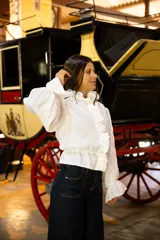 Camisa de lino blanca con cuello alto con volados, cierre frontal con botones ocultos y detalle de volados verticales. Presenta mangas largas abullonadas con puños acampanados y ajuste elástico en la cintura.