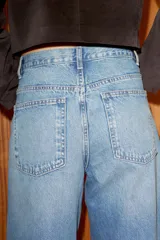 Jeans de tiro alto, color gris oscuro lavado, con pierna ancha extra larga (full length) y diseño de cintura cruzada asimétrica con cierre frontal de cremallera y botones metálicos.