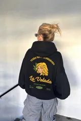 Canguro negro de corte oversize con capucha y bolsillo frontal. Presenta un diseño gráfico estampado en la espalda que simula un paquete de yerba mate, con la ilustración de una mujer, hojas y la inscripción 'La Volada'.