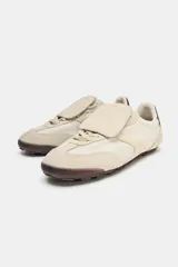 Championes deportivos de estilo minimalista en color beige, confeccionados en una combinación de materiales textiles y gamuza. Presentan una lengüeta superpuesta con cierre de gancho, punta redonda y un detalle de textura oscura en el talón. Suela plana de goma en tono marrón.