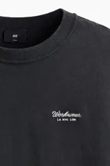 Remera de algodón de punto pesado en color negro con efecto lavado, cuello redondo acanalado, hombros caídos y pequeño estampado tipográfico en el pecho.