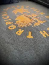 Hoodie gris oscuro con capucha, de calce estándar, con estampa en color amarillo en la espalda que presenta una palmera, un atardecer y la leyenda Tropical Sunset Palm Beach.