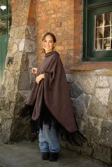 Poncho de tejido suave color marrón chocolate, con diseño amplio y relajado. Presenta detalles artesanales de costura en los bordes y en el bolsillo frontal, además de flecos largos en color negro en todo el ruedo.