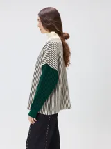 Pullover de tejido acanalado grueso con cuello alto y cierre frontal parcial. Presenta un diseño de bloques de color, con cuerpo en tono beige y mangas en contraste con rayas verticales en tonos rojizos. Incluye un detalle bordado en el pecho.