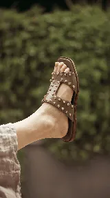 Sandalia baja de cuero tostado, con tiras anchas en forma de H adornadas con tachas semiesféricas doradas.