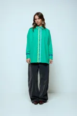Camisa azul de corte oversize, con cierre frontal de botones y detalles de cintas onduladas en colores contrastantes a lo largo de la botonadura y en los puños. Presenta cuello clásico y mangas largas.