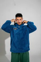 Canguro de textura tipo peluche en color azul, con cierre frontal parcial, cuello alto y bolsillo tipo canguro. Presenta un parche ovalado en el pecho con diseño gráfico.
