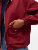 Campera tipo parka corta de color beige, con capucha ajustable con cordón, cierre frontal y grandes bolsillos frontales con solapa. Presenta puños elásticos fruncidos.