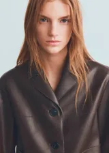Chaqueta de piel sintética en color marrón, de diseño recto, manga larga y cuello con solapa. Cuenta con cierre frontal de botones, dos bolsillos laterales y forro interior.