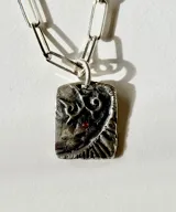 Collar con cadena de eslabones tipo clip y dije rectangular de plata con diseño texturizado, relieves abstractos y un pequeño cristal rojo incrustado.