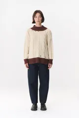 Buzo de tejido de punto con diseño de trenzas, color crema con detalles en marrón en cuello, puños y ruedo. Presenta líneas finas en contraste y pequeñas costuras decorativas en forma de cruz.