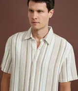 Camisa de hombre confeccionada en 100% algodón con textura, de manga corta y cuello inglés. Presenta un diseño de rayas verticales en tonos beige, blanco y marrón, con cierre frontal de botones.