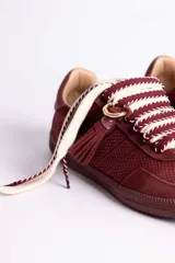 Zapatillas urbanas color tostado con textura geométrica en el cuerpo y detalles de gamuza en la puntera y laterales. Presentan un diseño distintivo con cordones anchos tejidos en tonos tostado y blanco, y un accesorio metálico con borla de gamuza en el empeine.