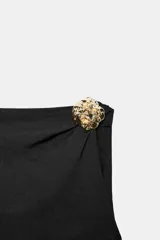 Body negro de manga corta con escote asimétrico, detalle de fornitura metálica dorada en relieve en el hombro y tejido drapeado en el torso. Cierre inferior con botones a presión.