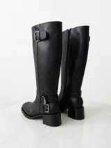 Bota de caña alta estilo biker, confeccionada en cuero negro. Presenta tres hebillas metálicas decorativas, cierre lateral y taco de madera.