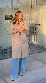 Trench coat color beige de corte clásico, con solapas amplias, doble botonadura frontal, cinturón ajustable con hebilla y trabillas en los puños.