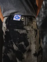Pantalón unisex de denim con diseño amplio y silueta barrel, inspirado en la bombacha de campo y el pantalón sastrero. Cuenta con lavado irregular efecto tie-dye en tonos negro y gris, pinzas delanteras y bolsillos laterales.