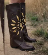 Bota de caña alta color marrón oscuro, confeccionada en cuero, con diseño de sol aplicado en tonos dorados y detalles de tachas metálicas.