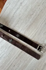 Cinturón de cuero vacuno color chocolate con apliques metálicos en forma de estrella y tachas circulares. Cuenta con hebilla metálica plateada.