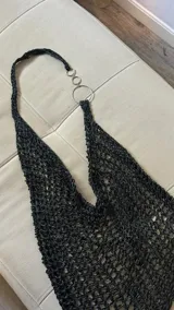 Top tejido a mano en hilo negro con textura de red abierta, escote halter con tiras que se unen a anillos metálicos plateados. Posee una etiqueta de cuero marrón con el nombre de la marca en la base.